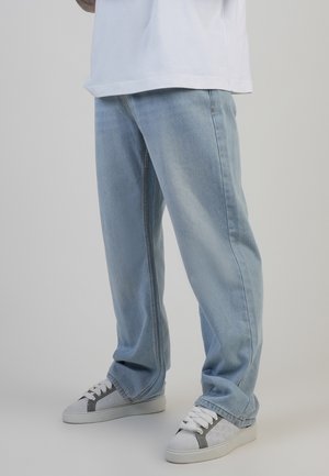 Person trägt lockere hellblaue Jeans und weiße Turnschuhe mit grauen Akzenten, steht vor einem schlichten hellen Hintergrund.