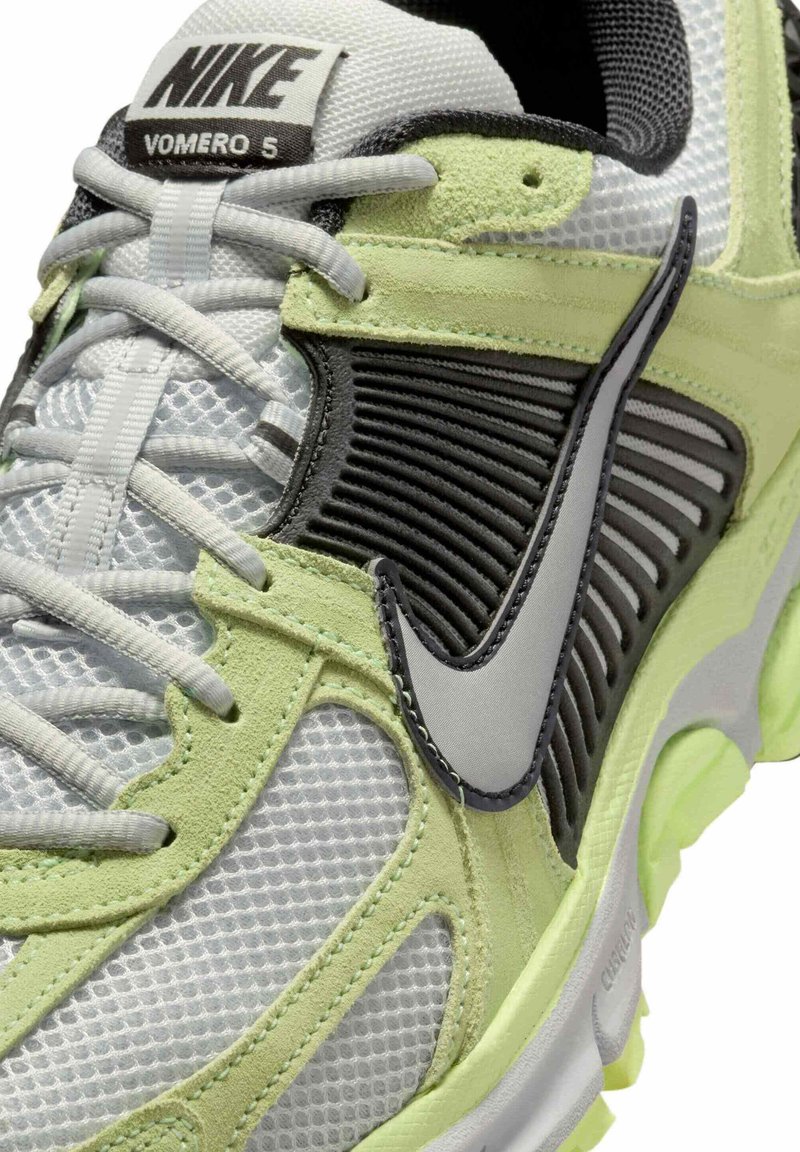 La zapatilla de correr Nike Vomero 5 presenta una parte superior de malla en gris claro con detalles en negro y verde, paneles texturizados y una suela acolchada.