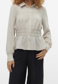 Blusa button-up di colore beige chiaro, con colletto, tessuto testurizzato, vita arricciata e maniche lunghe. Indossata con pantaloni scuri e larghi.