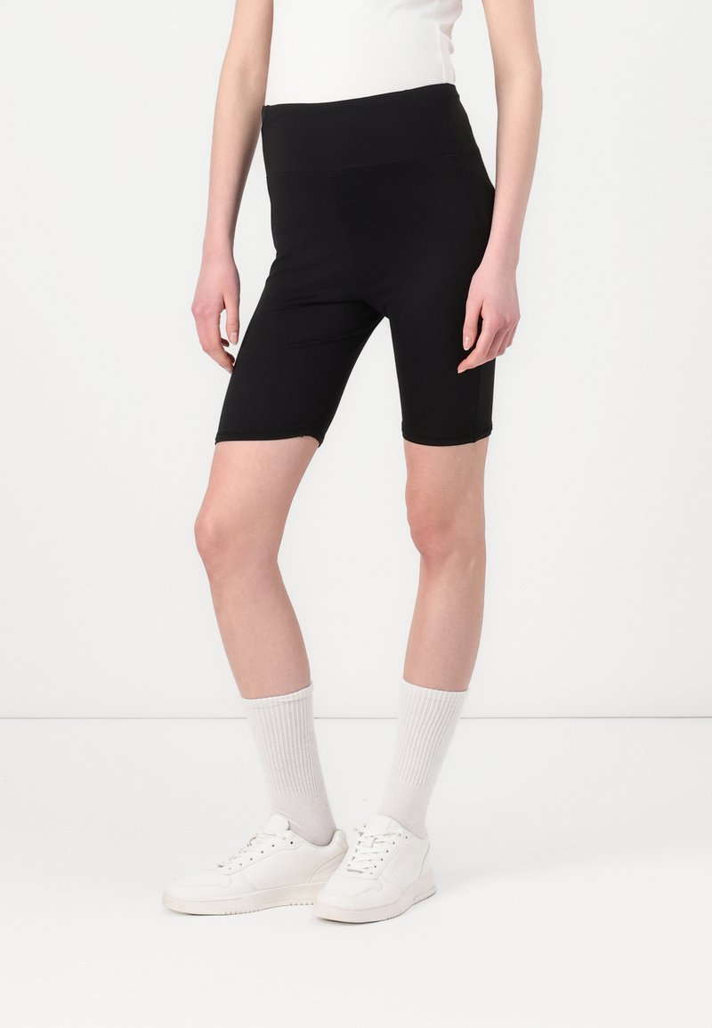 Zwarte hoogwaist fietsbroek gemaakt van rekbaar materiaal, met een gladde textuur en een aansluitende silhouette, gecombineerd met witte sneakers en sokken.