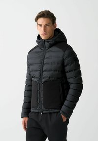 Schwarze Pufferjacke mit Kapuze, horizontal gestepptem Muster und einfarbigen schwarzen Akzenten an Taschen und Schultern. Reißverschluss vorne.