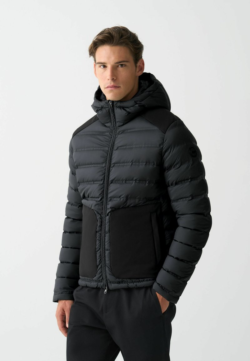 Schwarze Pufferjacke mit Kapuze, horizontal gestepptem Muster und einfarbigen schwarzen Akzenten an Taschen und Schultern. Reißverschluss vorne.