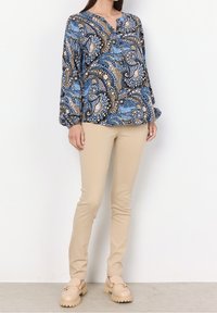 Blouse à manches longues bleue à motif cachemire avec des motifs dorés et blancs, associée à un pantalon beige ajusté et des chaussures beiges épaisses.