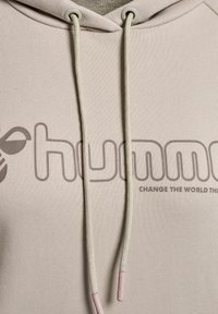 Haut à capuche beige avec une texture côtelée, affichant un grand logo gris et le texte "CHANGE THE WORLD" dans une police simple. Comprend des cordons de serrage de couleur claire.
