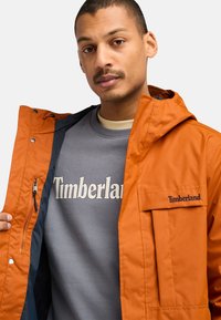 Orange vattenavvisande jacka med huva, som har en ficka med dragkedja och Timberland-logotyp. Bärs över en grå sweatshirt med textdesign.