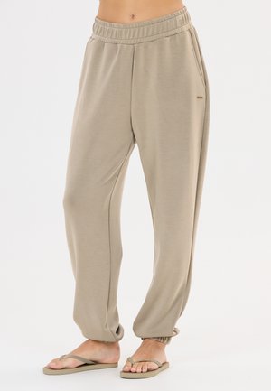 Losse beige sweatpants met elastische tailleband en boorden, gedragen met bijpassende beige slippers bij een staand persoon.