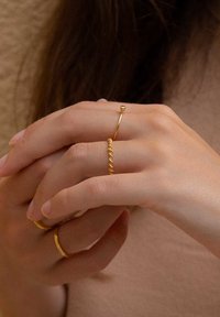 Goldringe an Händen, einschließlich eines gedrehten Bands und eines dünnen Rings mit einem kleinen Perlendetail. Glatte Textur, polierte Oberfläche. Neutraler Hintergrund.