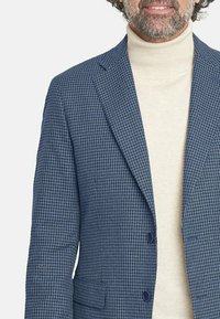 Blazer azul con patrón de espiga, con solapas con muesca y dos botones, combinado con un suéter de cuello alto color crema por debajo.