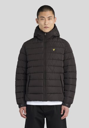 Mann trägt schwarze gesteppte Daunenjacke mit Kapuze, Reißverschluss vorne, Seitentaschen und kleinem gelben Vogel-Logo auf der Brust vor einfarbigem grauem Hintergrund.