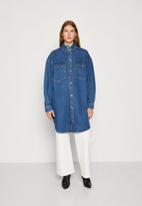 Denim srajčna obleka, temno modra z dvema sprednjima žepoma, zapiranje na gumb, dolgi rokavi in splošno sproščen kroj, v kombinaciji z belimi širokimi hlačami.