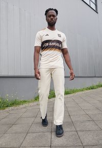 Maglia atletica beige chiaro con dettagli neri e dorati, che presenta la scritta "LOS ANGELES" e strisce laterali nere, abbinata a pantaloni beige e scarpe nere.