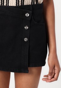 Svart denimkjol med sidknäppning med silverknappar, framficka och rak passform. Slät textur och kort längd.