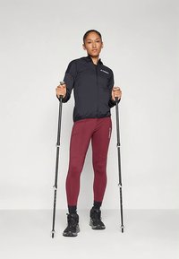 Γυναίκα με μαύρη ζακέτα και μπορντό κολάν στέκεται κρατώντας μαύρες μπαστούνες πεζοπορίας σε ουδέτερο φόντο.