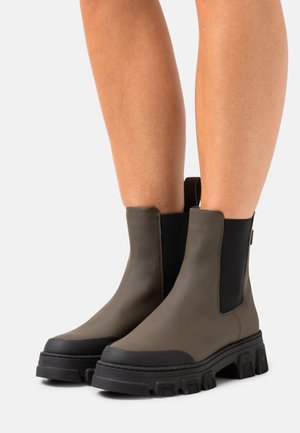 Bottes de cheville en caoutchouc vert olive avec une semelle noire épaisse, des panneaux latéraux élastiques et une languette pour un enfilage facile. Fini lisse avec un design robuste.