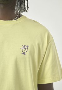 Gelbes Kurzarm-T-Shirt aus Baumwolle, mit einem lila Vogelmotiv auf der linken Brustseite und einer glatten Textur.