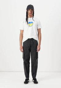 Hvid T-shirt med blåt og gult logo, sorte cargo-bukser med sidelommer, sorte sneakers med tre hvide striber og grå kasket.