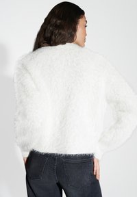 Femme de dos portant un pull blanc en peluche à manches longues et un jean noir taille haute, les mains sur les hanches, devant un fond uni.