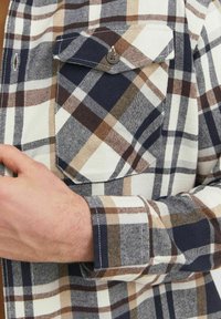 Camisa de franela a cuadros en navy, blanco y marrón. Presenta un bolsillo en el pecho, cierre de botones y mangas largas con un patrón de cuadros.