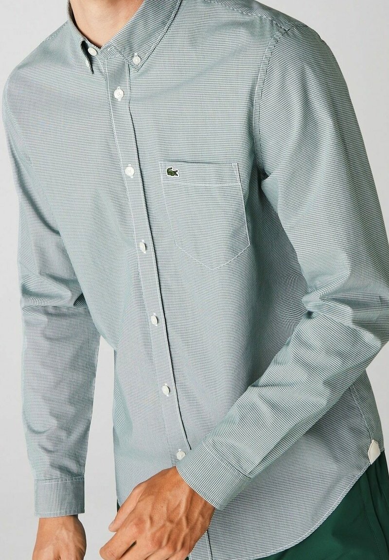 Chemise à manches longues à carreaux verts et blancs avec boutons, dotée d'une poche avant et d'un logo brodé sur la poitrine, fabriquée en tissu léger.