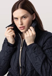 Veste matelassée marine avec un col montant, fermeture éclair à l'avant et poignets élastiques. Elle présente un motif texturé et une taille avec cordon de serrage.