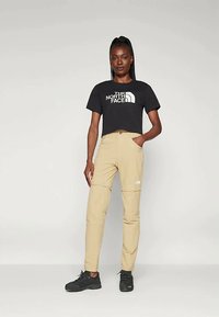 Juoda trumpa palaidinė su "The North Face" logotipu, derinama su smėlio spalvos kelnių su užtrauktukais prie kelių ir juodais sportbačiais.