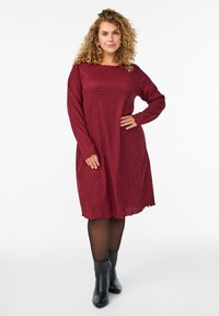 Femme aux cheveux bouclés portant une robe rouge à manches longues avec de petits pois, des collants noirs et des bottines noires, se tenant avec une main sur la hanche.