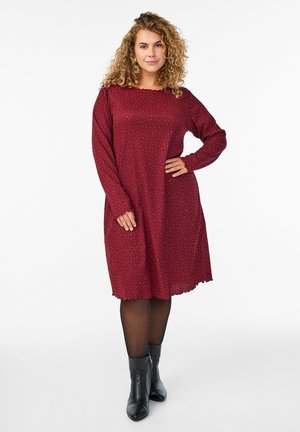 Vrouw met krullend haar, gekleed in een rood jurk met lange mouwen en kleine stippen, zwarte panty en zwarte enkellaarsjes, die staat met één hand op haar heup.