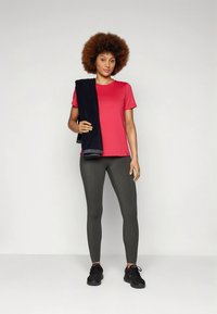 Rød atletisk skjorte med korte ermer, grå tights og svarte joggesko; holder et mørkt håndkle over skulderen; ensfarget bakgrunn.