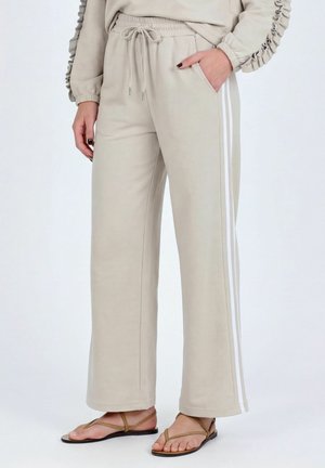 Pantalon de survêtement beige à jambes larges avec des bandes latérales blanches, taille élastique, cordon de serrage, porté avec des sandales plates beiges à lanières.