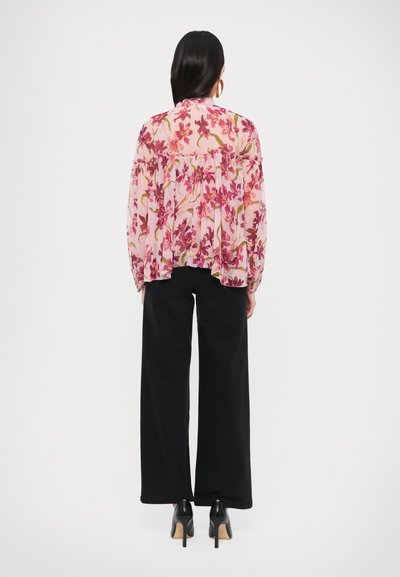 Blouse fleurie rose clair avec des motifs violets et verts, dotée d'un ourlet à volants. Associée à un pantalon noir taille haute à jambes larges et des talons.