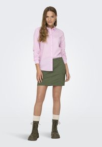 Camicia button-up a strisce verticali di colore rosa chiaro abbinata a una minigonna verde khaki. Stivali marroni con lacci e risvolti color crema.
