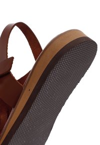Sandale en cuir marron avec une semelle en caoutchouc noir texturé, design plat, sangle réglable et semelle intermédiaire de couleur naturelle.