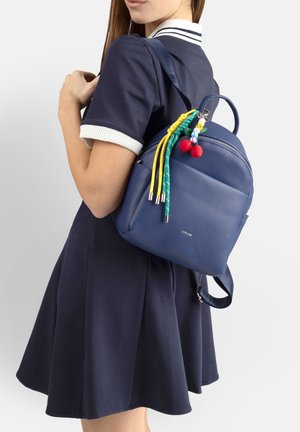 Donna in abito blu navy che indossa uno zaino in pelle blu con nappine intrecciate colorate e pompon rossi su sfondo bianco.