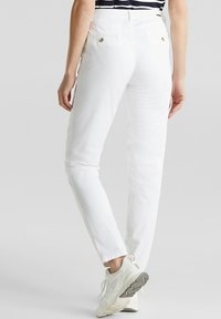 Pantalon en coton blanc avec une coupe slim, poches arrière, et boutons dorés, assorti à des chaussures de sport gris clair.