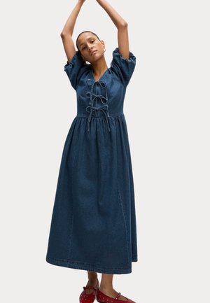 Denim midi kjole med puffærmer, v-hals og snørehåndtag foran. Har en glat tekstur og en løs pasform for komfort. Røde sko med prikket mønster.