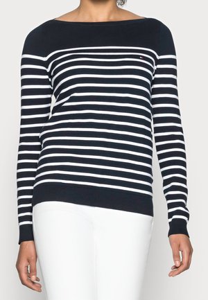 Person wearing a navy blue and white horizontal striped long-sleeve sweater and white pants, torso visible, neutral background.