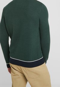 Mörkgrön, stickad tröja med ett strukturerat mönster, som har en marinteblå ribbad nederkant och en vit accentlinje. Bärs över beige byxor.