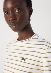 T-shirt bianca con righe orizzontali verdi e gialle, con un piccolo logo ricamato a forma di coccodrillo sul petto e un colletto rotondo.