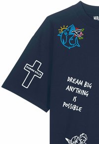 T-shirt bleu marine avec un requin brodé mangeant des fleurs, croix blanche avec « Karma Kills » sur la manche, et texte « Rêvez grand, tout est possible. »