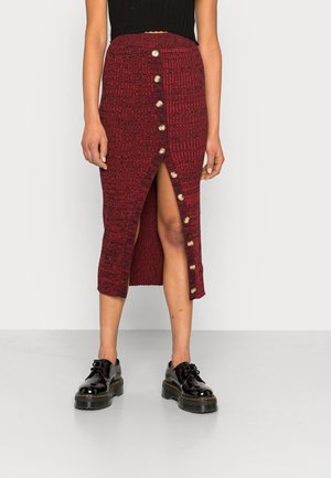 Femme portant une jupe tricotée rouge longueur genou avec des boutons sur le devant et une fente à l'avant, associée à des chaussures noires épaisses à lacets et un haut noir.