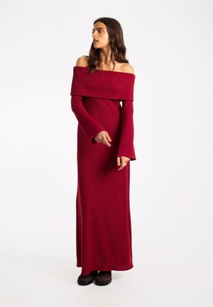 Lungo abito rosso off-the-shoulder realizzato in tessuto morbido, caratterizzato da maniche lunghe con polsini a campana e una silhouette aderente.
