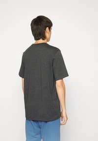 Male wearing a dark gray short-sleeve t-shirt and blue pants, standing facing a plain white wall with hands relaxed by sides.