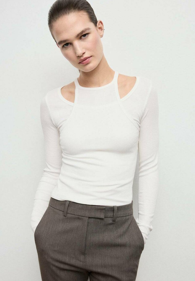Top bianco a maniche lunghe con dettagli spalle scoperte, realizzato in un materiale a coste. Abbinato a pantaloni grigi su misura, con una vestibilità morbida.