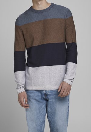 Mężczyzna w długim swetrze z dzianiny z poziomymi blokami kolorów: niebieskim, brązowym, granatowym i szarym, w połączeniu z jasnoniebieskimi dżinsami.