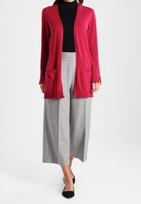 Cárdigan de punto magenta con abertura frontal, dos bolsillos delanteros, usado sobre un cuello de tortuga negro, combinado con pantalones de pierna amplia gris claro y zapatos negros.