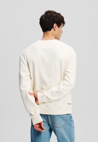 Crème-kleurige sweatshirt met lange mouwen, ribgebreide boorden en zoom. De achterkant toont zachte stof en een klein logo nabij de zoom. G gedragen met lichte denim jeans.