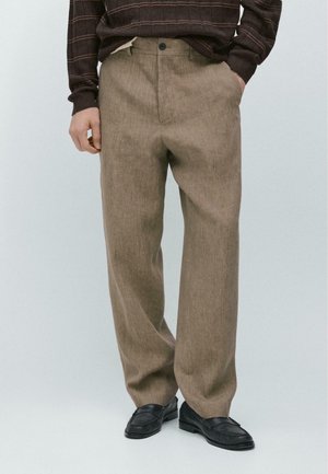 Homme portant un pantalon beige ample, des mocassins noirs et un pull rayé foncé, une main dans la poche.