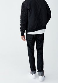 Veste bomber noire avec poignets côtelés, associée à un jean en denim foncé avec une couture rouge subtile, et des baskets blanches à rayures noires.