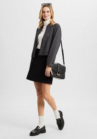 Graue, verkürzte Jacke, weißer Pullover mit Rollkragen, schwarzer Rock, schwarze Loafers und beigefarbene Socken. Hält eine schwarze Handtasche mit goldenen Hardware-Akzenten.