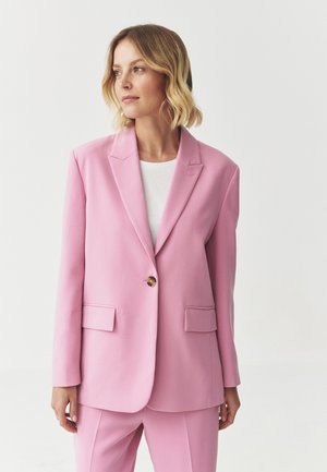Light pink tailored blazer featuring a single brown button, notched lapels, and two front pockets, paired with a white top.